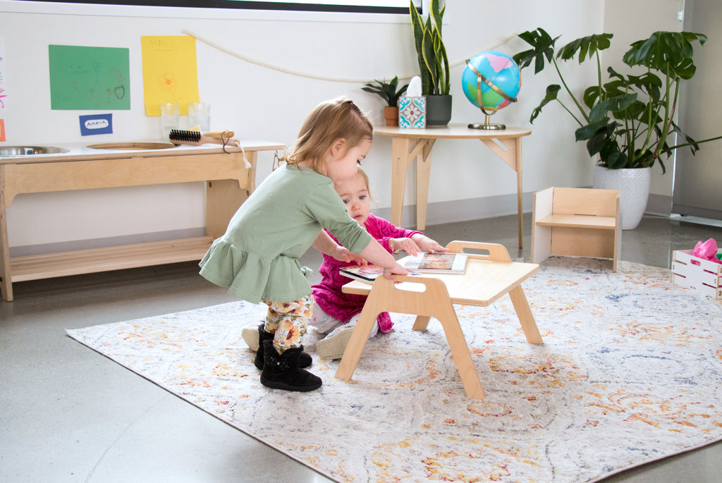 Two toddlers reading books on a floor table in a Montessori learning environment.